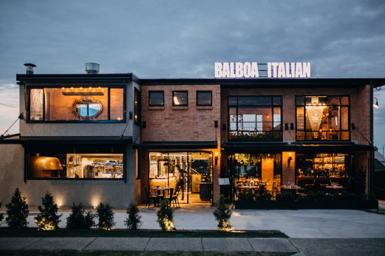 Exterior of Balboa Italian, one of the best restaurants in Palm Beach, featuring a warm, industrial-chic design with glowing interior lights and modern rustic appeal at dusk.