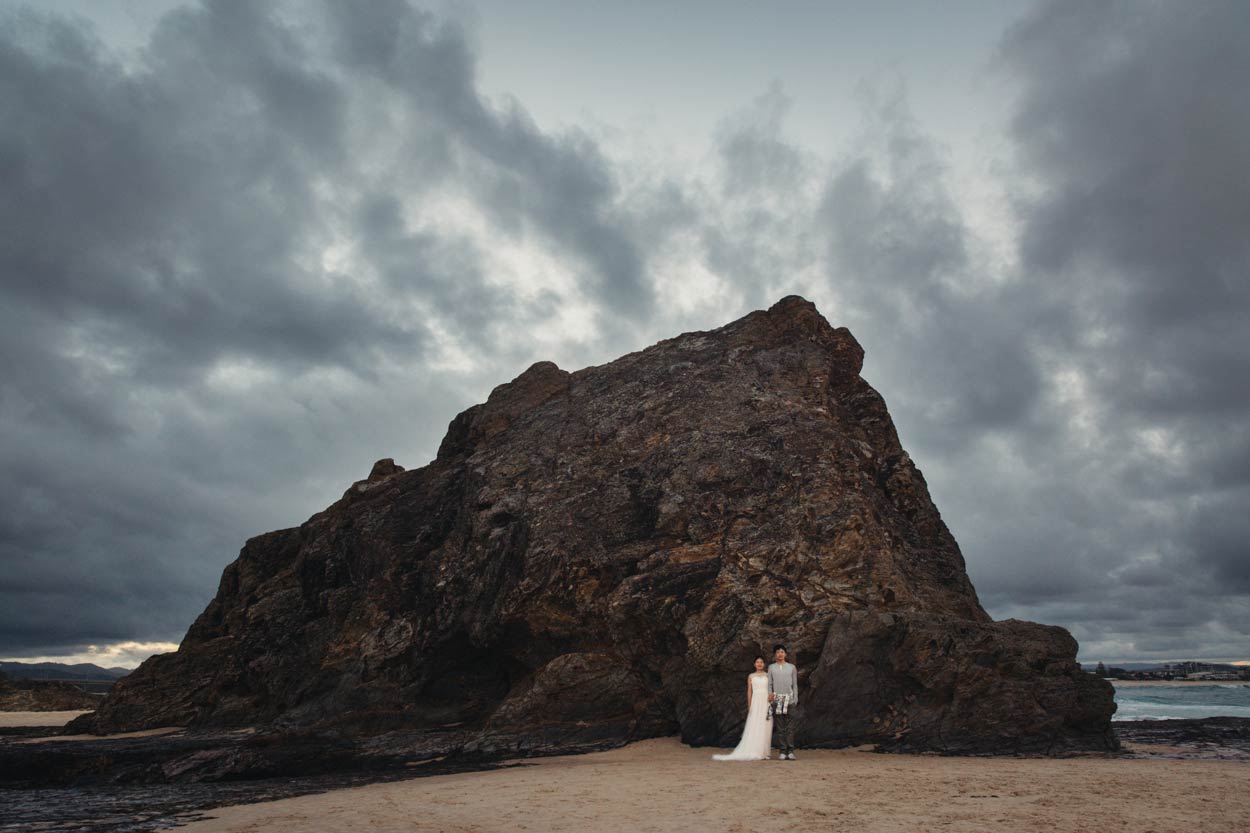 Newlyweds standing at the base of dramatic Currumbin Rock under moody skies — one of the best places in Gold Coast for unforgettable coastal scenery and photo-worthy moments.