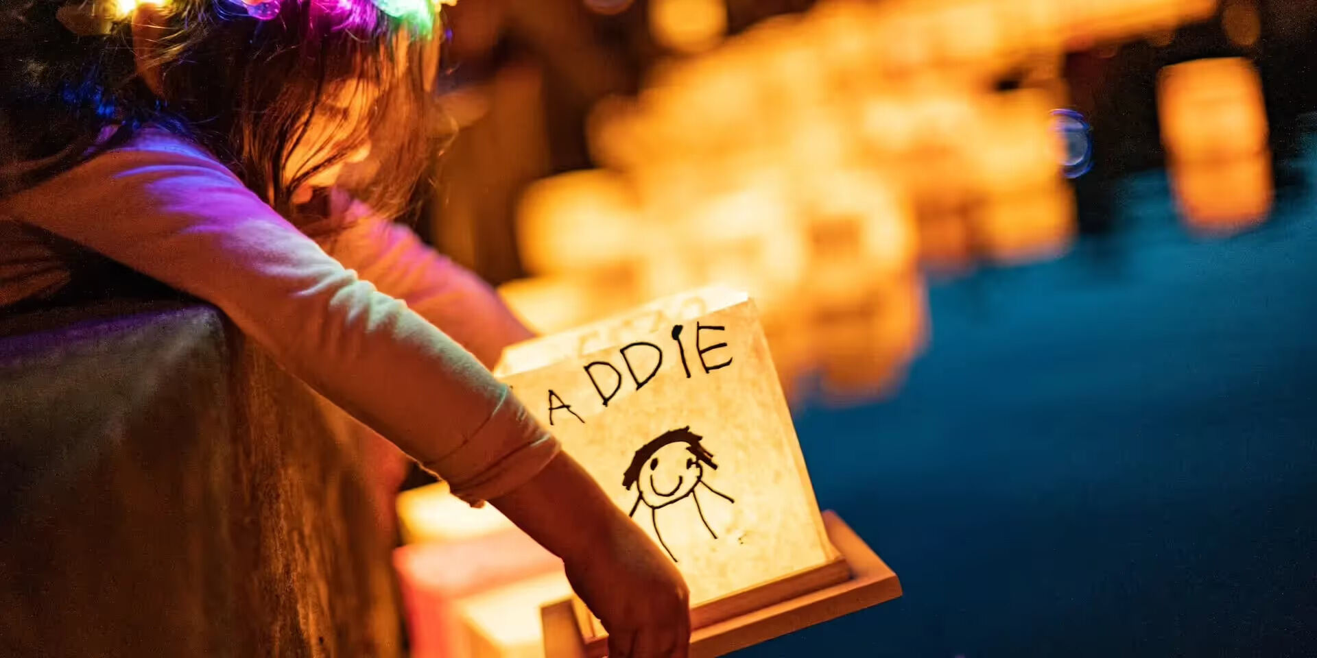 gold-coast-entertainment Gold coast entertainment at a water lantern festival, with a child holding a glowing personalised lantern beside the water at night